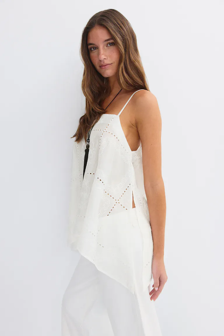 Hvit, brodert camisole med tynne spaghettistropper, asymmetrisk nederkant og sidespalte, kombinert med hvite bukser med vidde ben og et langt, svart anhengskjede.