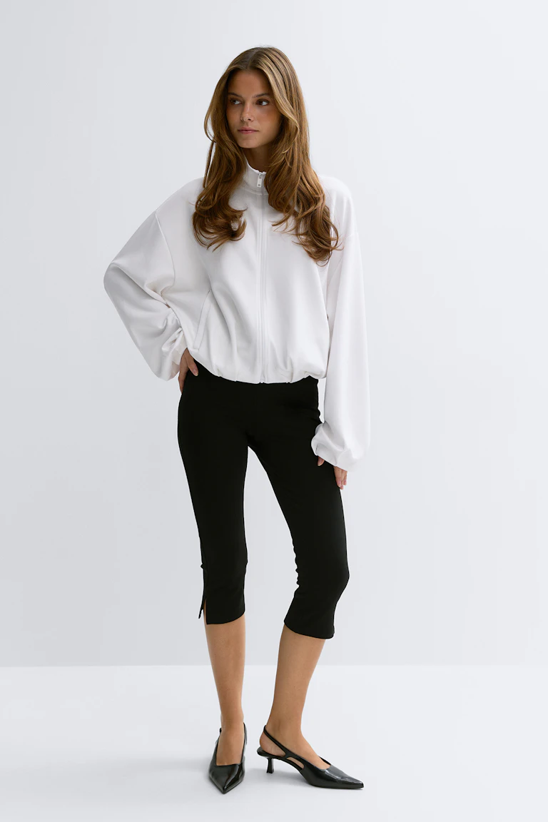 Valkoinen vetoketjullinen ylisuuri takki, jossa korkea kaulus ja pitkät hihat; mustat kapeat caprihousut, joissa on pienet sivuhalkiot; mustat teräväkärkiset kitten-korkoiset slingback-kengät.