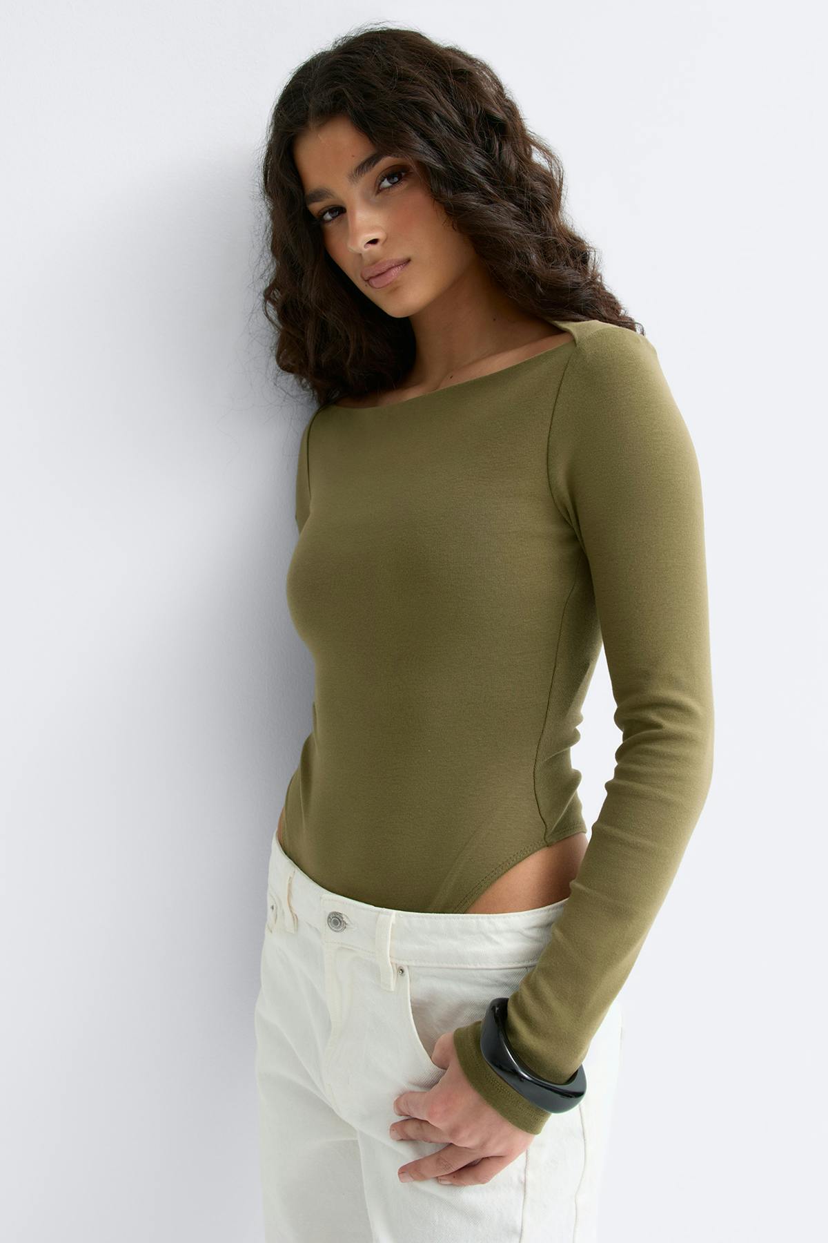 Oliivinvihreä pitkähihainen sovitettu bodysuit, jossa on veneen pääntie, yhdistettynä korkeavyötäröisiin valkoisiin farkkuihin ja mustaan paksuiseen rannerenkaaseen.