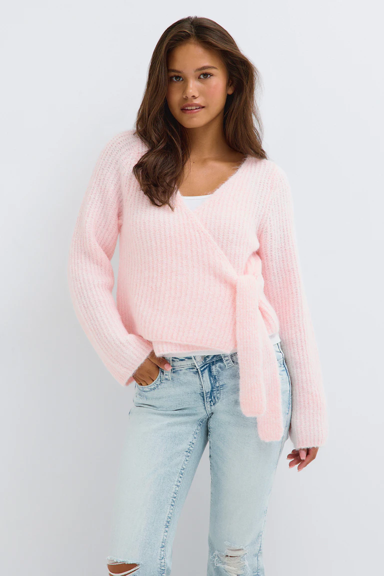 Ein hellrosa Wickelpullover über einem weißen Oberteil, dazu eine hellgewaschene Jeans mit Rissen.