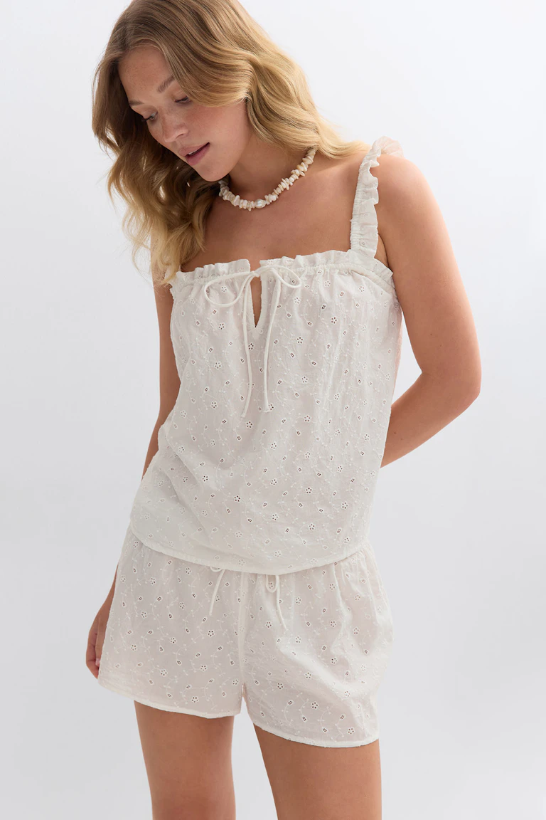 Valkoinen eyelet-koristeinen camisole, jossa on röyhelöiset olkaimet ja solmio edessä, yhdistettynä yhteensopiviin kiristysnauhalla varustettuihin eyelet-shortseihin.