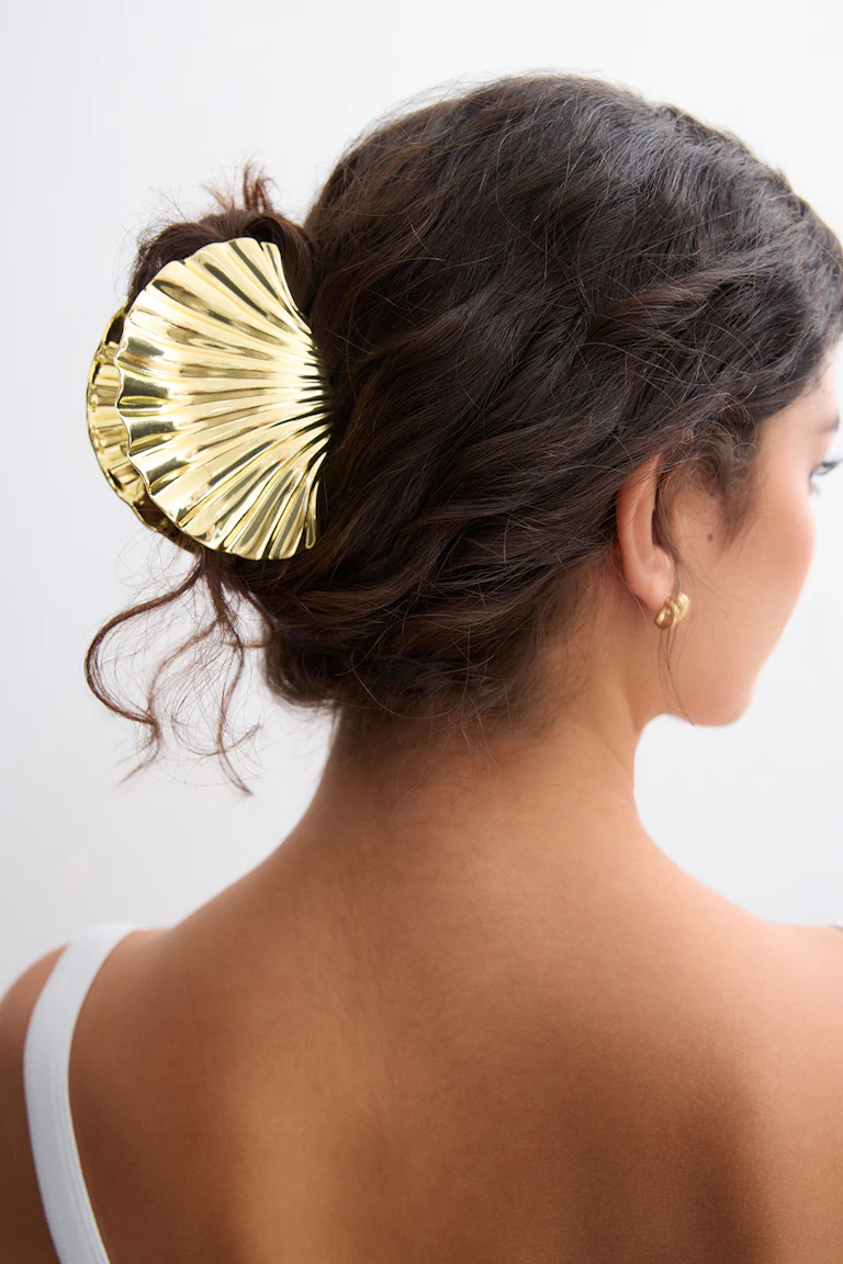 Pince à cheveux dorée en forme de coquillage fixant un chignon bas, petites boucles d'oreilles à nœuds dorés et bretelles d'un haut sans manches blanc.