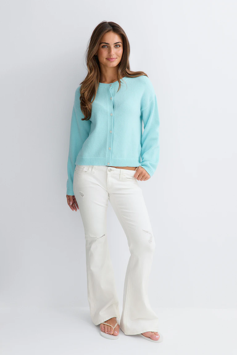 Un cardigan couleur aqua clair, à manches longues, col rond et boutons sur le devant, associé à un jean évasé blanc délavé et à des sandales à lanières blanches.