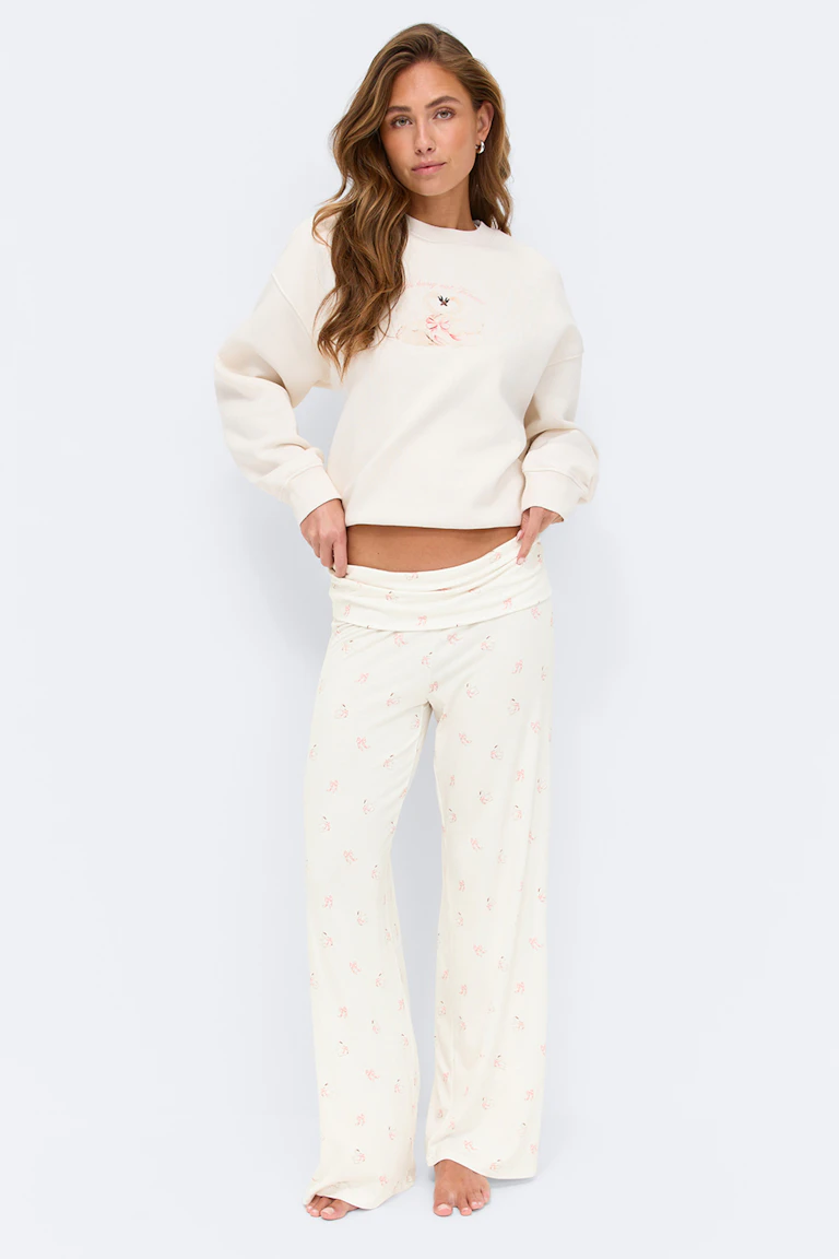 Crèmekleurig sweatshirt met subtiel bloemmotief op de borst en ontspannen lange mouwen, gecombineerd met een bijpassende crèmekleurige pyjamabroek met wijde pijpen met een kleine roze bloemenprint en omgeslagen tailleband; blootsvoets.