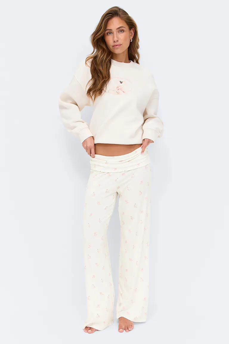 Crèmekleurig sweatshirt met subtiel bloemmotief op de borst en ontspannen lange mouwen, gecombineerd met een bijpassende crèmekleurige pyjamabroek met wijde pijpen met een kleine roze bloemenprint en omgeslagen tailleband; blootsvoets.