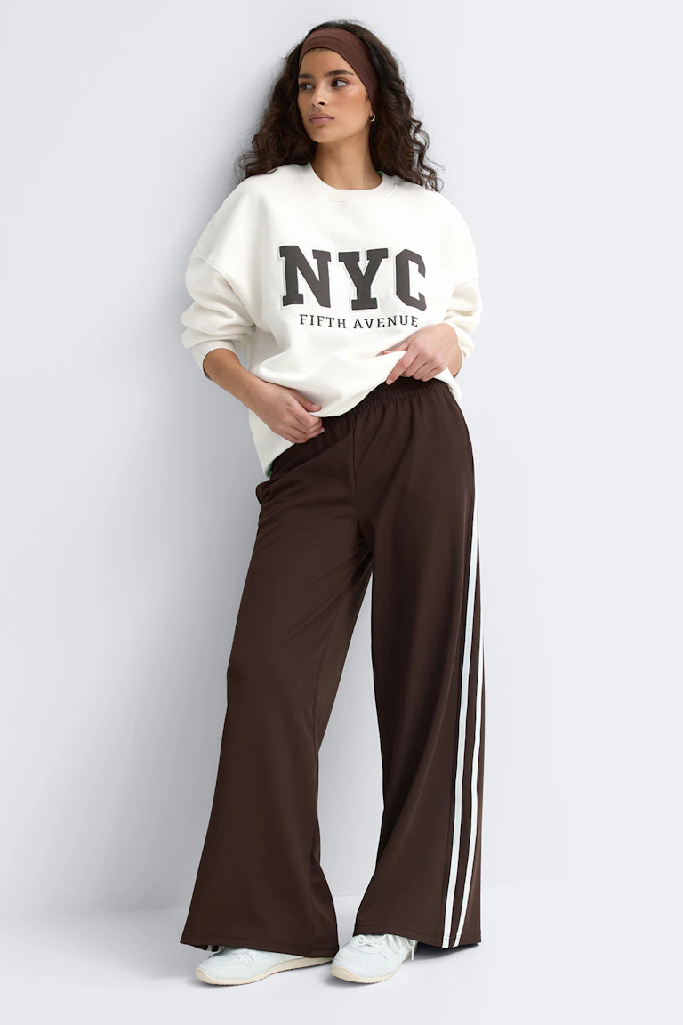 Das Model trägt ein übergroßes cremefarbenes Sweatshirt mit dem Aufdruck "NYC Fifth Avenue", eine braune Trainingshose mit weitem Bein und weißen Seitenstreifen, weiße Turnschuhe und ein braunes Stirnband.