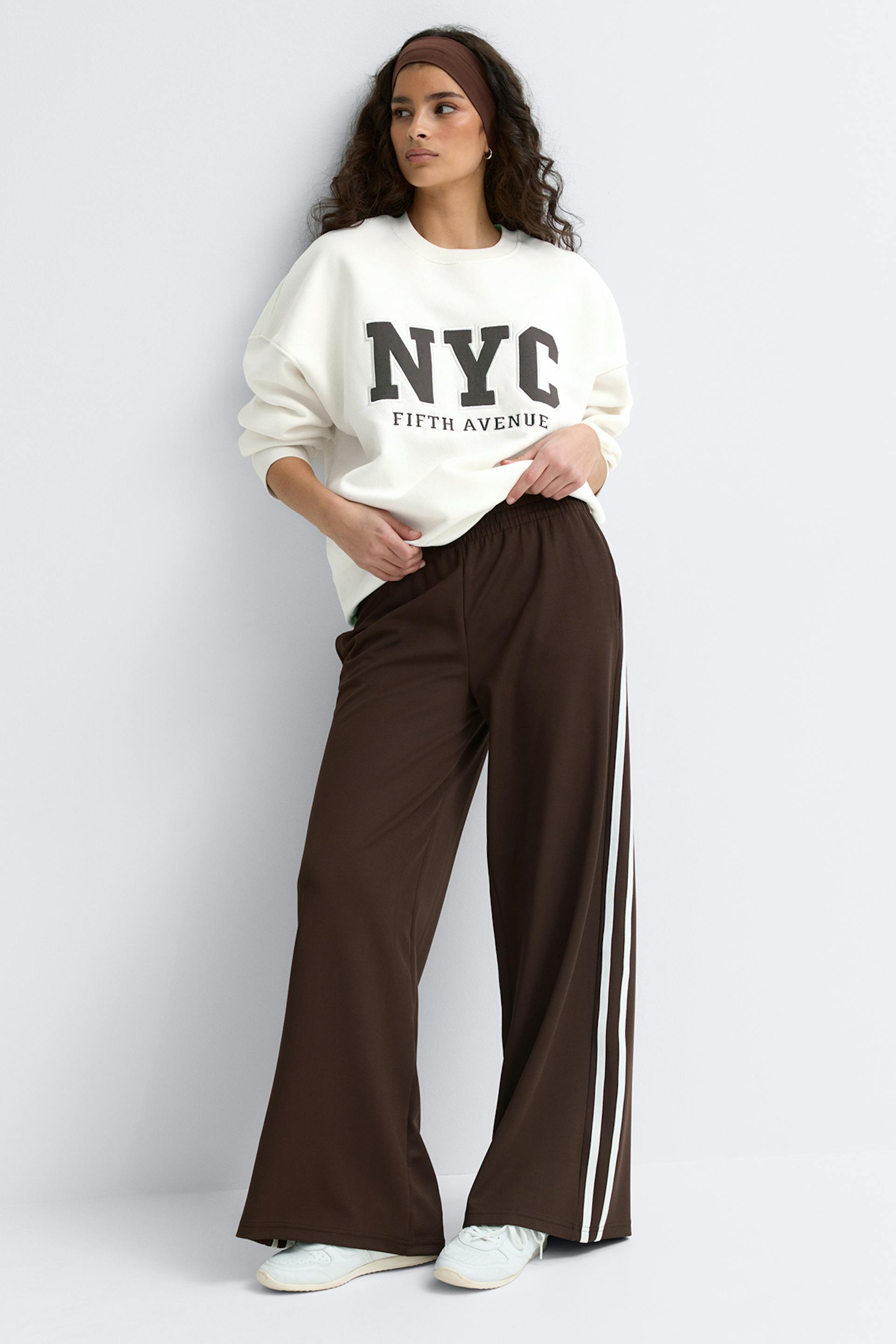 Mallilla on yllään ylimitoitettu luonnonvalkoinen collegepaita, jossa on teksti "NYC Fifth Avenue", ruskeat leveälahkeiset verkkarit, joissa on valkoiset sivuraidat, valkoiset lenkkarit ja ruskea pääpanta.