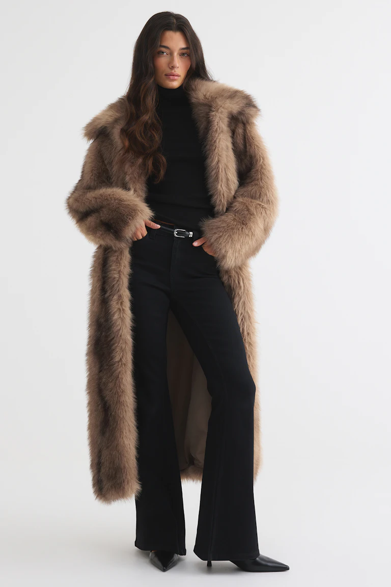 Een lange taupe jas van imitatiebont over een zwarte coltrui, een zwarte broek met wijde pijpen, een zwarte riem en zwarte hakken met spitse neus.