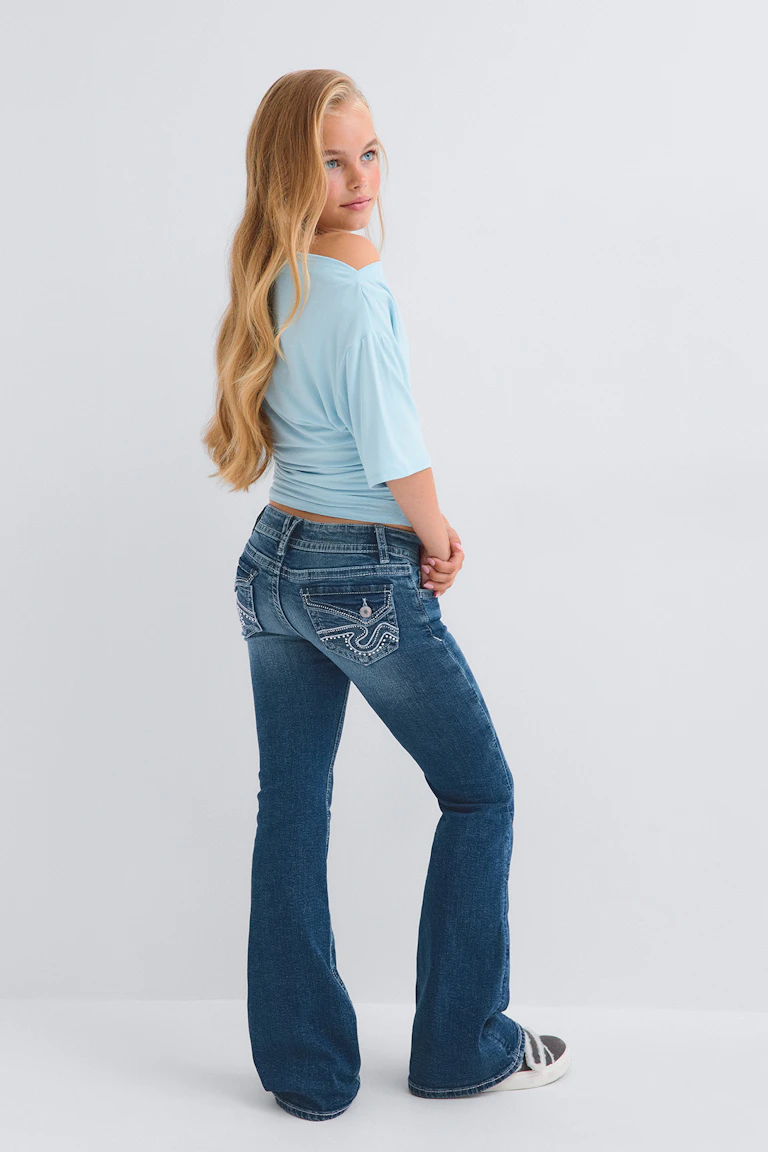 Hellblaues, schulterfreies Oberteil mit dunkelblauen, ausgestellt geschnittenen Jeans und grau-weißen Sneakers.