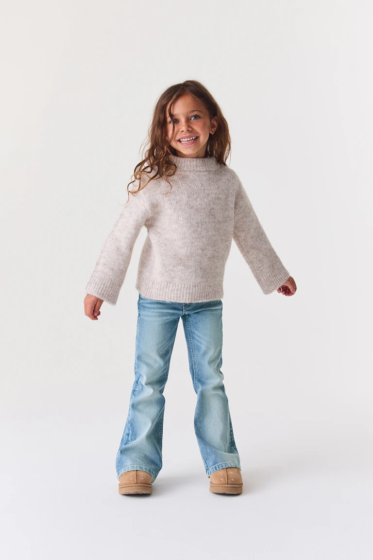Ein hellbeiger, flauschiger Pullover mit hohem Kragen, hellblaue Schlagjeans und hellbraune Stiefel.