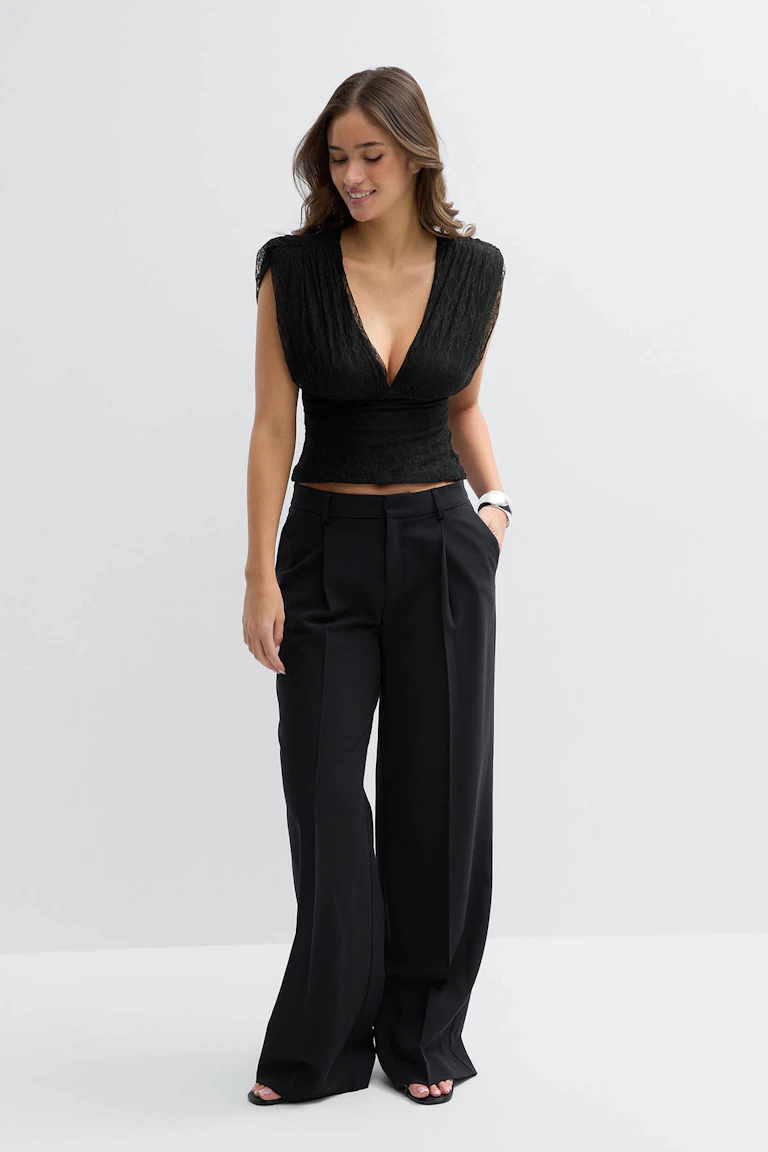 Schwarzes ärmelloses Oberteil mit tiefem V-Ausschnitt aus Spitze, gepaart mit einer schwarzen Hose mit hoher Taille und weitem Bein und schwarzen offenen Absätzen.