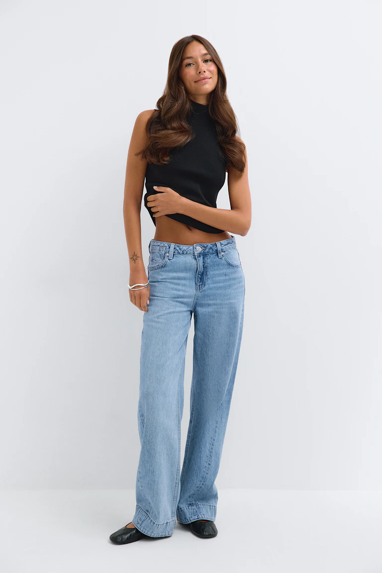 Zwarte mouwloze top met hoge kraag, lichtblauwe jeans met wijde pijpen en zwarte ballerina’s.
