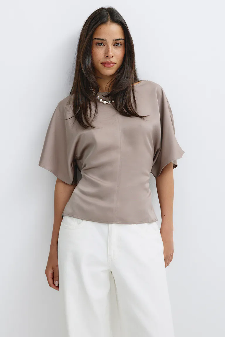 Seidenähnliche, taupefarbene Kurzarmbluse mit Kimono-Ärmeln und einer Mittelnaht, kombiniert mit einer weißen Hose mit hoher Taille und weitem Bein sowie einer silbernen Perlenkette.