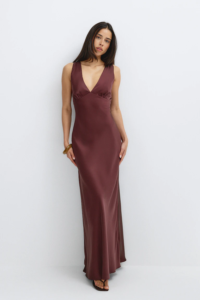 Robe nuisette sans manches en satin bordeaux, à encolure en V profond et longueur au sol, associée à des sandales noires à fines lanières entre les orteils et à un seul bracelet en résine ambrée.