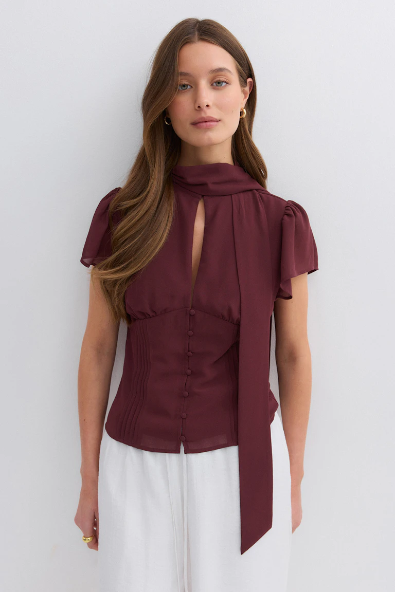Een bordeauxrode blouse met korte mouwen, een keyhole voorkant, een met stof bedekt button-down lijfje en een lange nekband, gecombineerd met een witte broek met wijde pijpen.