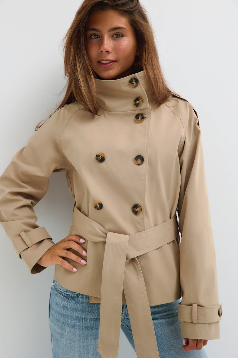 Beiger Trenchcoat mit doppelter Knopfleiste und breitem Revers, darüber ein weißes, kurzes Spitzen-Top mit Lochmuster und gewelltem Saum, dazu eine blaue Jeans in mittlerer Waschung.