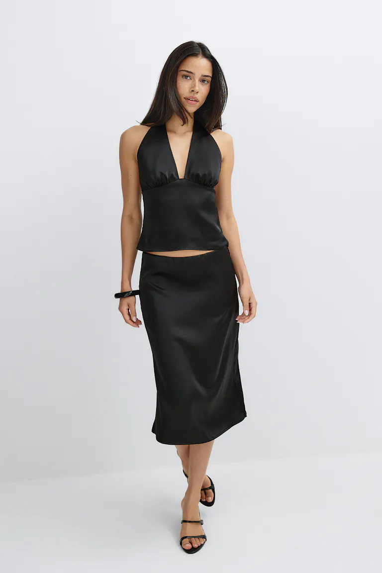 Ein schwarzes Satin-Haltertop mit tiefem V-Ausschnitt, kombiniert mit einem passenden schwarzen Satin-Midirock, schwarzen Riemchensandalen und einem schwarzen Armreif.