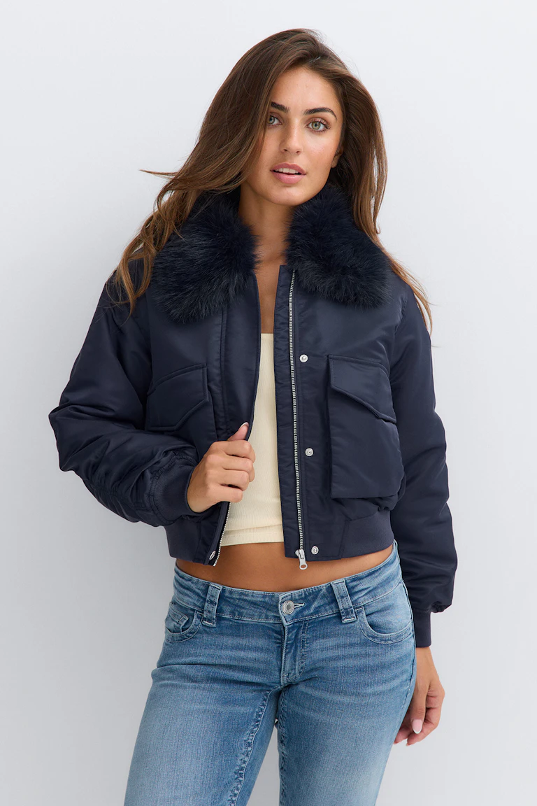 Une veste bomber bleu marine dotée d'un col en fausse fourrure sombre et moelleuse, de poches à rabat et d'une fermeture à glissière et à boutons-pression sur le devant, portée par-dessus un débardeur court couleur crème, le tout associé à un jean bleu délavé moyen.