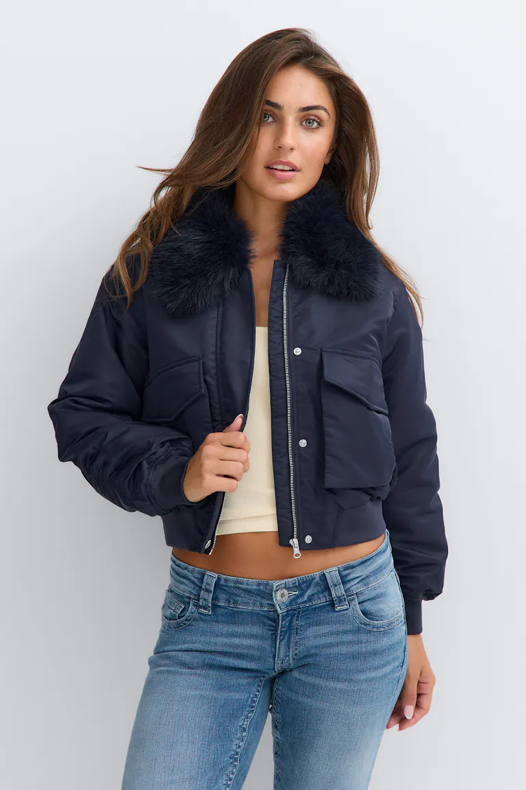 Navy bomber jacket with a plush dark faux‑fur collar, flap pockets and zip‑and‑snap front over a cream cropped tank top, paired with mid‑wash blue jeans.