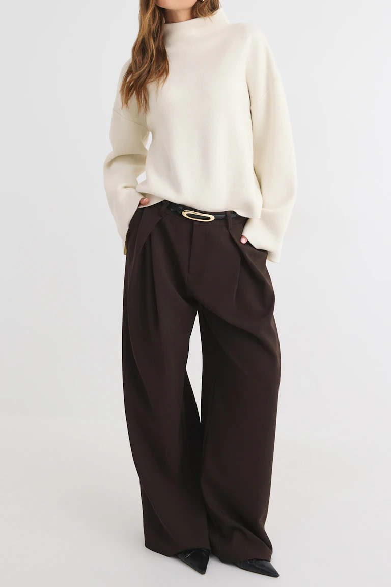 Cremefarbener, weit geschnittener Pullover mit hohem Kragen, dazu eine dunkelbraune Hose mit weitem Bein, ein schwarzer Gürtel mit goldener Schnalle und schwarze spitze Schuhe.
