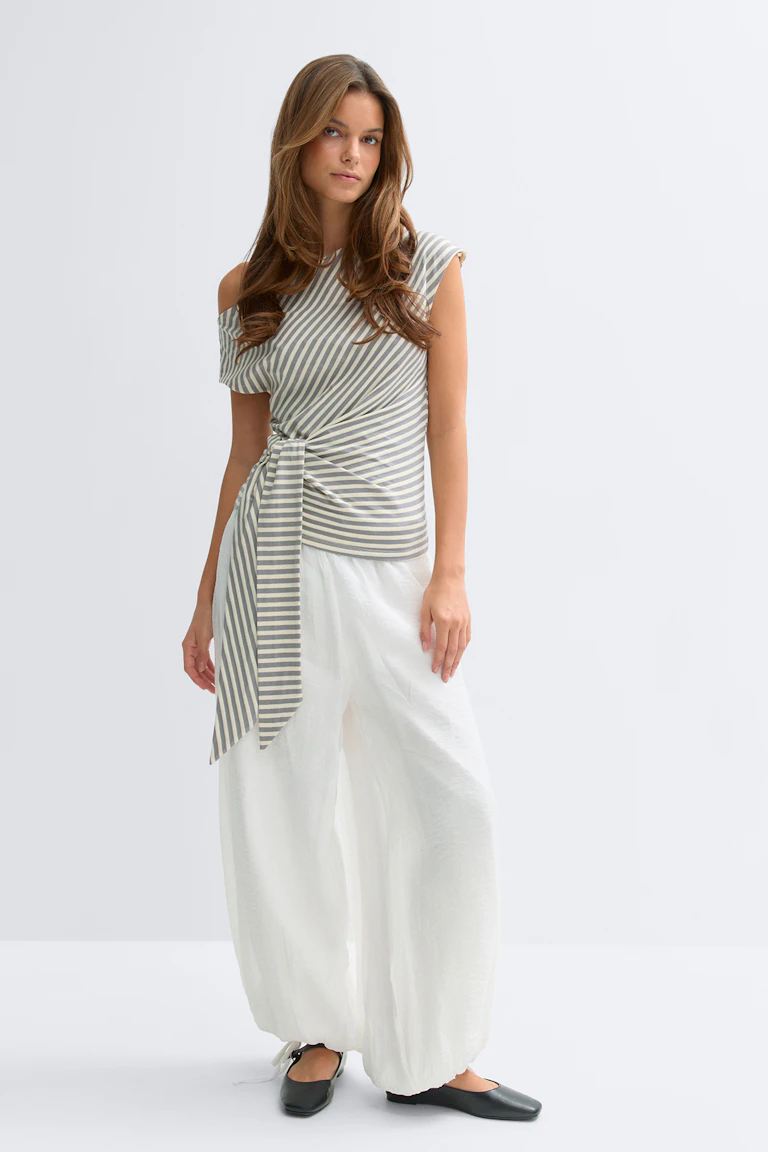 Grau-weiß gestreiftes, asymmetrisches One-Shoulder-Top mit seitlichem Knoten und langer Schärpe; weiße, geraffte Hose mit weitem Bein; schwarze Ballerinas.