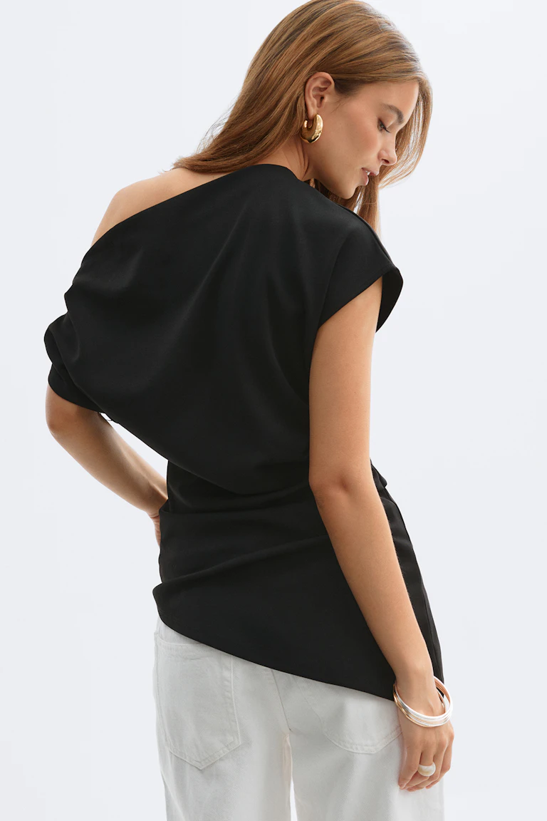 Top asymétrique noir drapé à une épaule associé à un pantalon blanc, accessoirisé de grandes boucles d'oreilles dorées et de bracelets métalliques superposés.