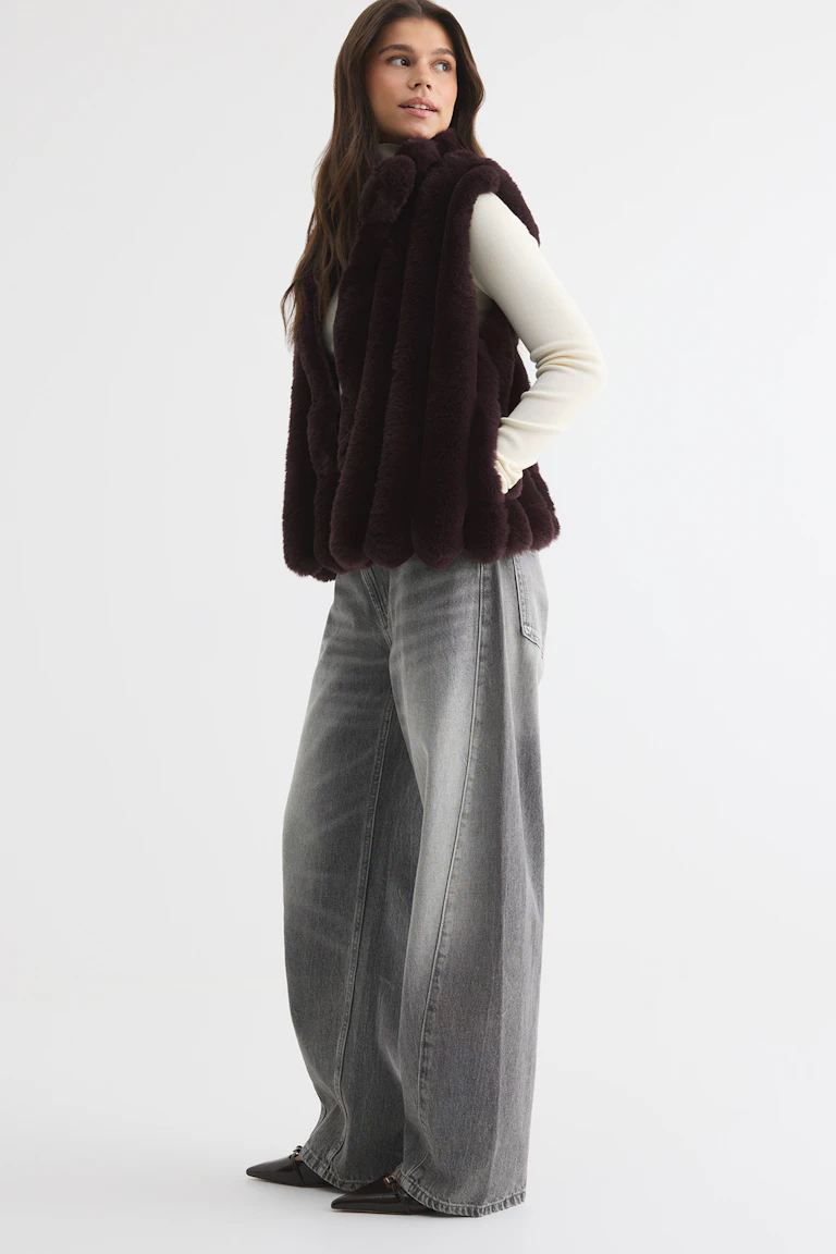 Eine dunkelbraune Kunstfellweste über einem elfenbeinfarbenen Langarm-Oberteil, dazu eine graue Jeans mit weitem Bein und schwarze spitze Ballerinas.