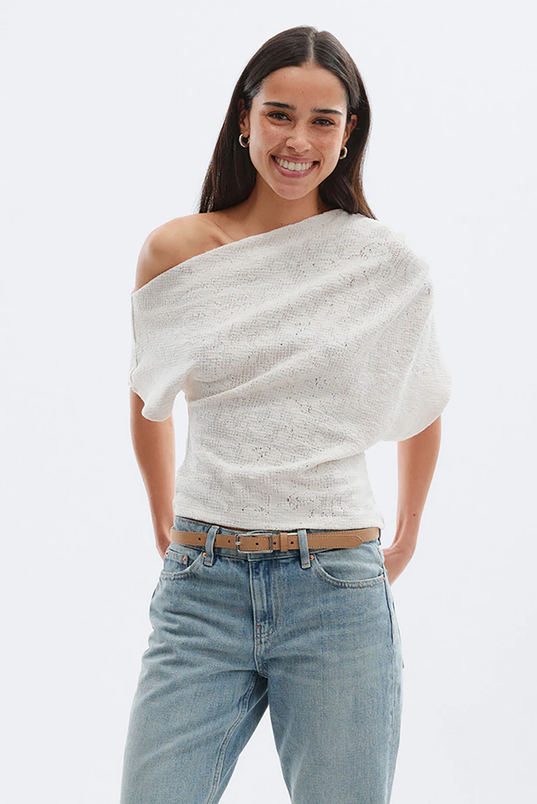 Gebroken witte gebreide top met één schouder en lichtgekleurde jeans met hoge taille, bruine leren riem en bruine leren schoenen met spitse teen.