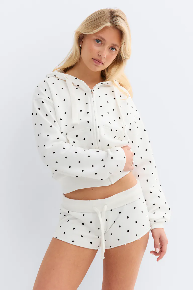 Weißer Kapuzenpullover mit Reißverschluss, Kapuze, Kordelzug und schwarzen Tupfen, kombiniert mit einer passenden weißen Lounge-Shorts mit elastischem Kordelzug und Tupfen.