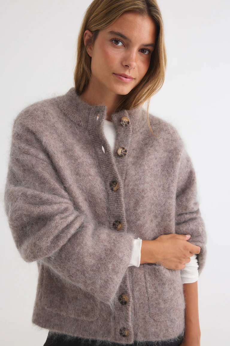 Eine taupefarbene, flauschige Strickjacke mit Knopfleiste, die über einem weißen Oberteil getragen wird.
