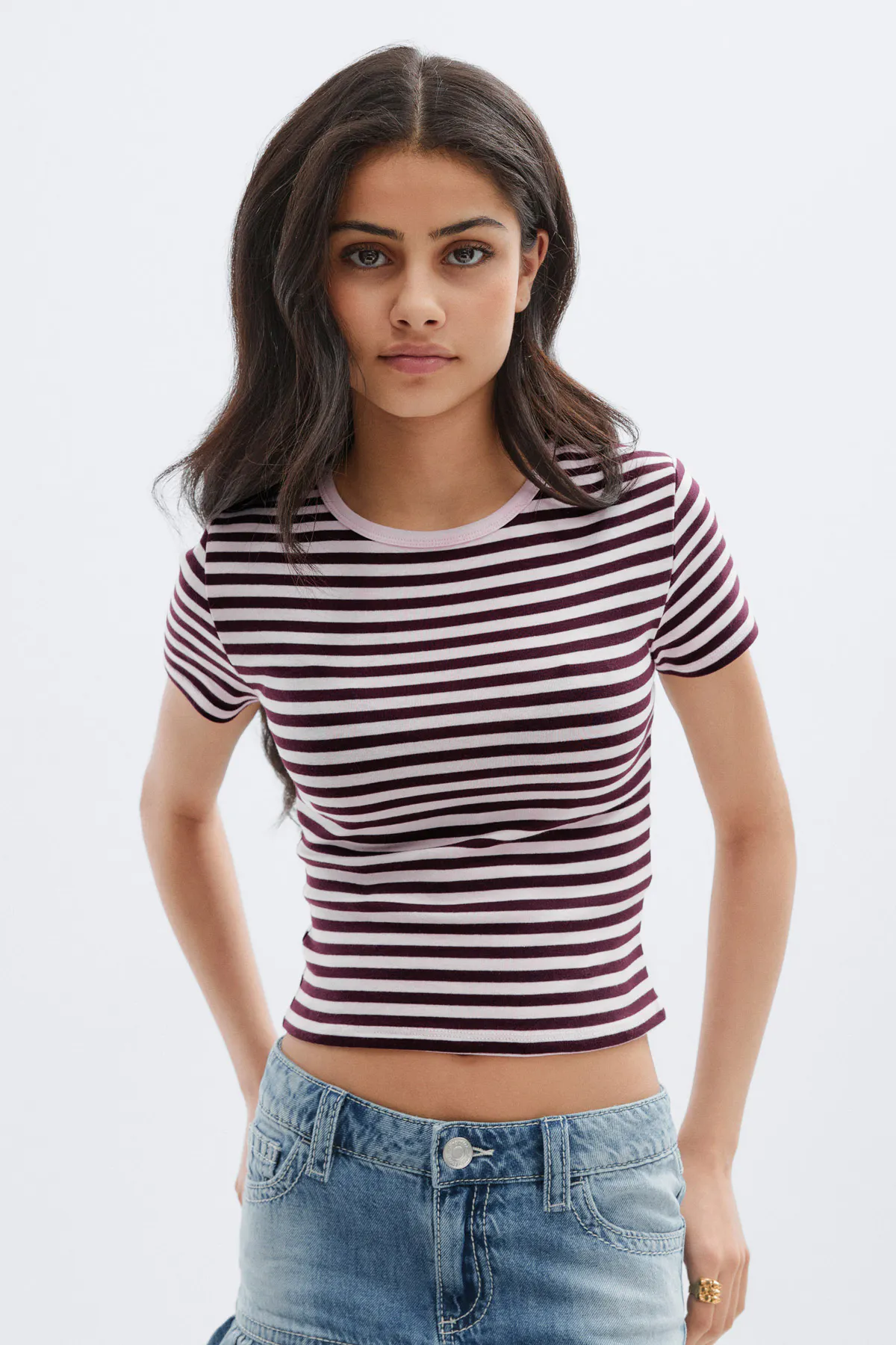 Istuva, lyhennetty lyhythihainen T-paita, jossa on viininpunaisia ja vaaleanpunaisia vaakaraitoja, yhdistettynä matalavyötäröisiin, kevyesti pestyihin farkkuihin.