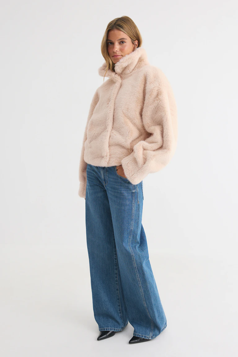 En modell bär en ljusrosa jacka i fuskpäls, blå jeans med vida ben och svarta skor med spetsig tå.
