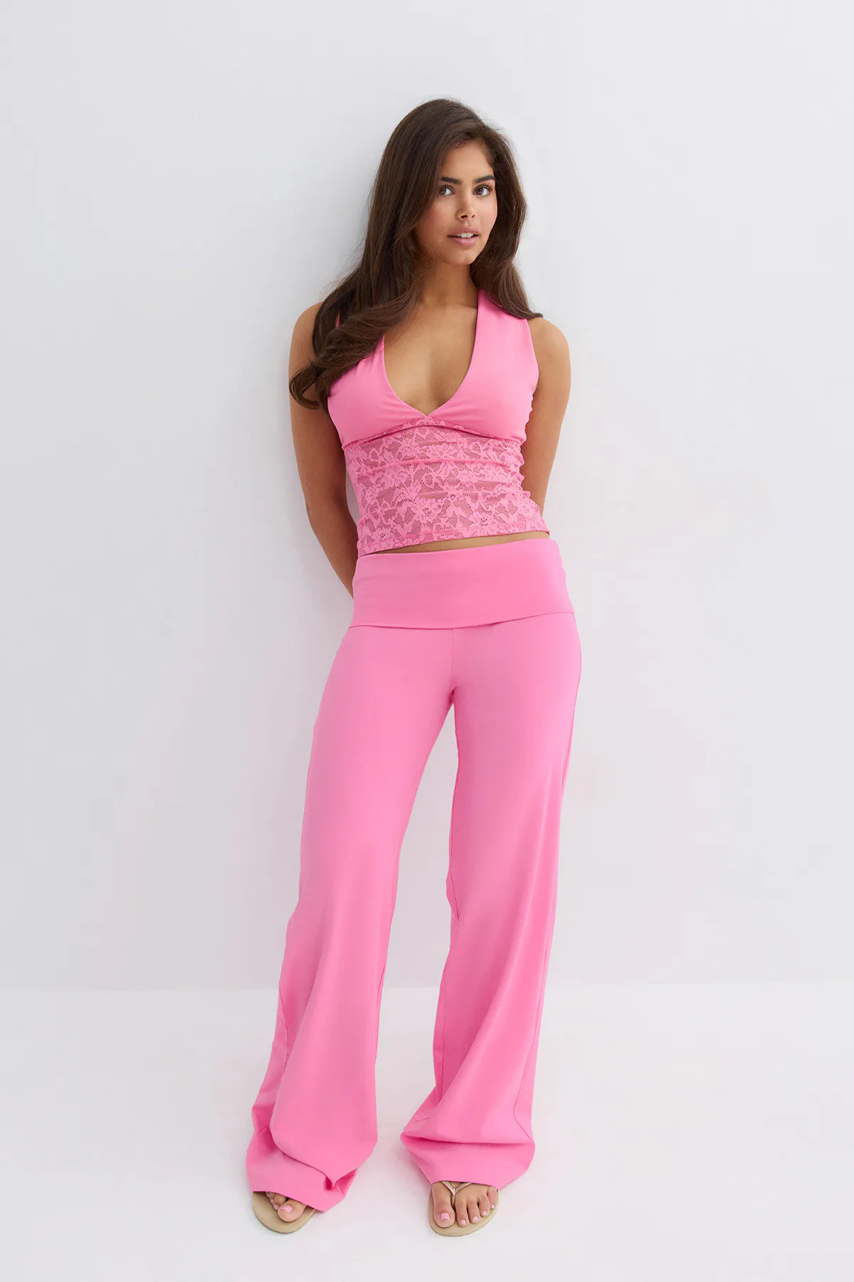 Rosafarbenes, ärmelloses Crop-Top mit V-Ausschnitt und Spitzeneinsatz in der Mitte, kombiniert mit einer passenden, hochtaillierten Hose mit weitem rosa Bein und beigen Zehensandalen.