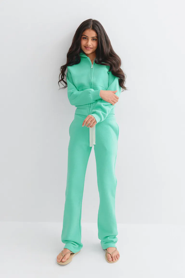 Personne portant un sweat à capuche zippé menthe-turquoise avec une poche kangourou sur le devant et un pantalon de survêtement à cordon de serrage assorti, associé à des tongs beiges.