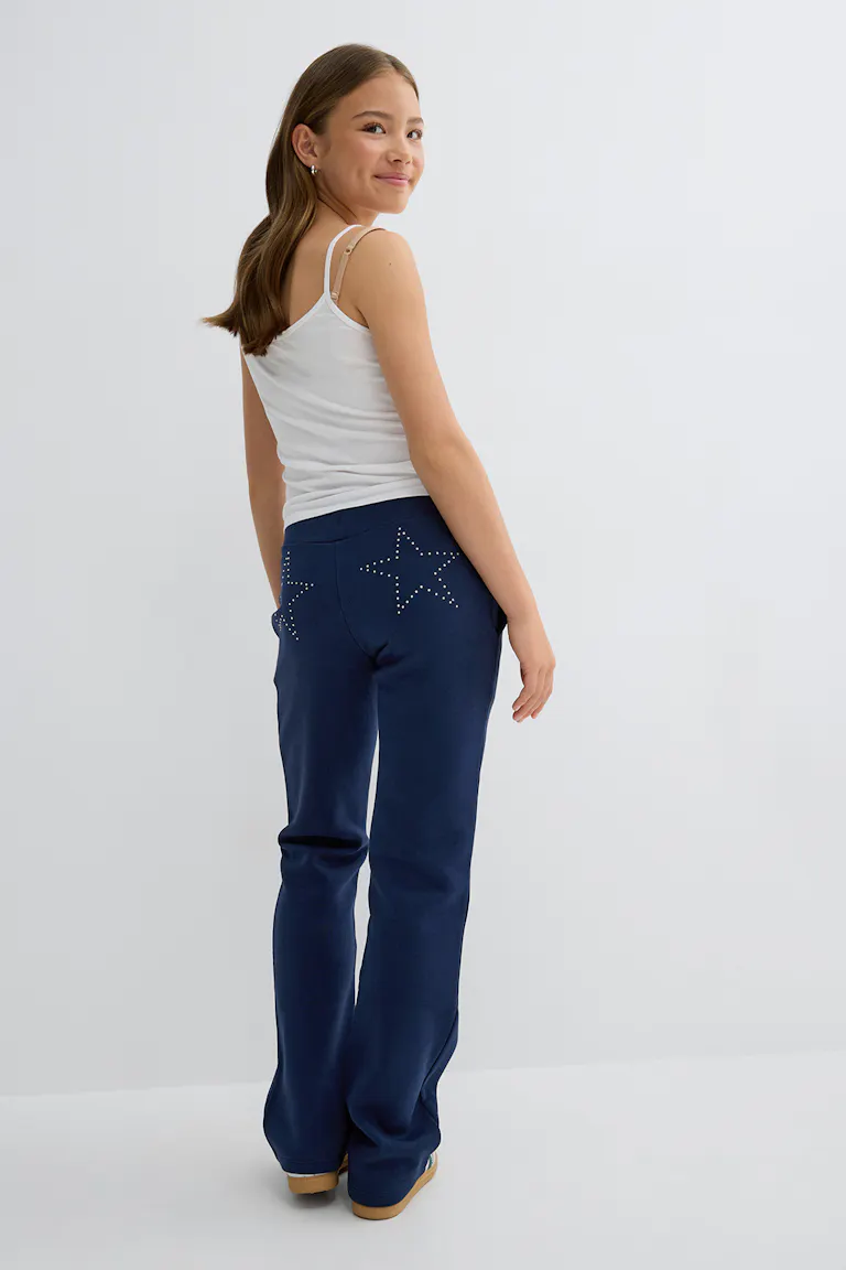 White fitted camisole with thin straps paired with navy sweatpants featuring studded star motifs on the back pockets, and tan slip-on shoes.