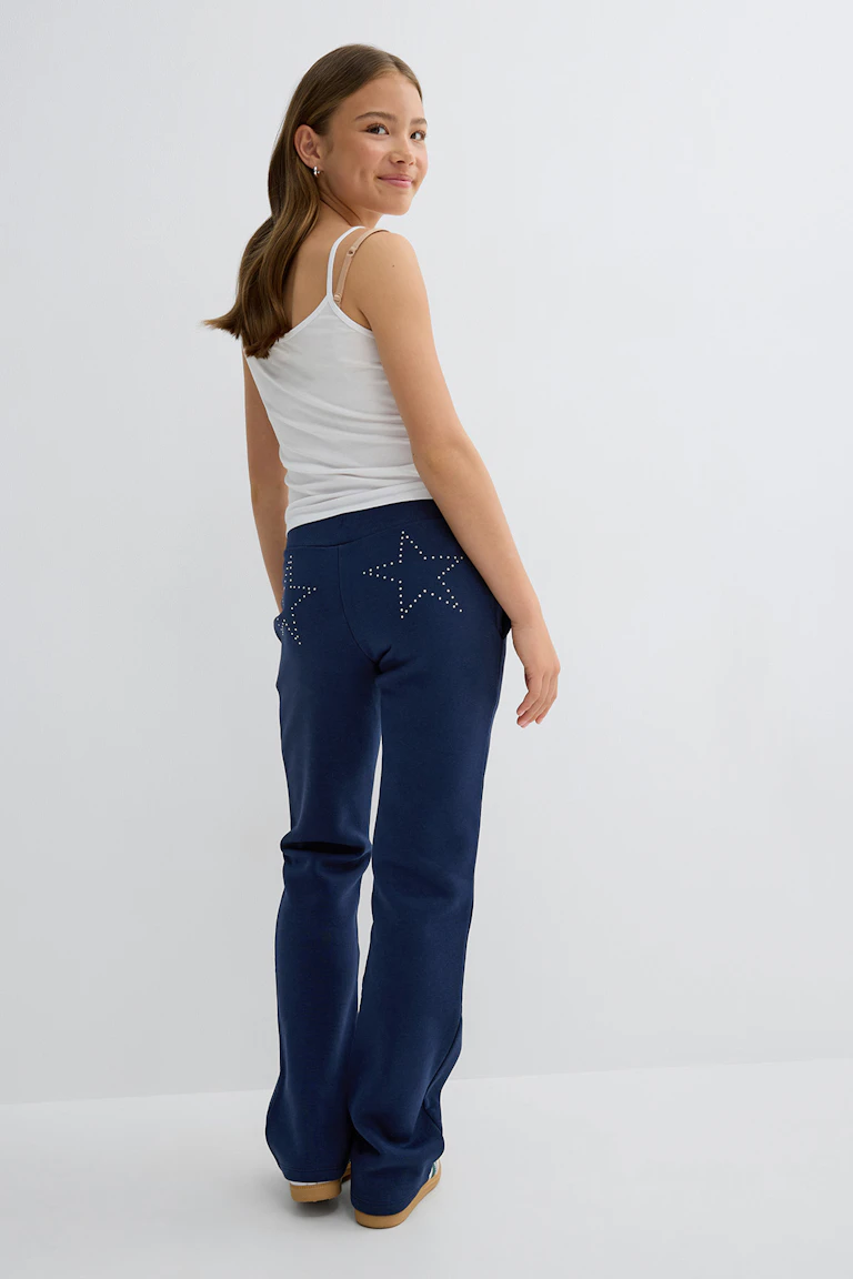 Caraco blanc ajusté à fines bretelles associé à un pantalon de survêtement bleu marine avec des motifs étoilés sur les poches arrière, et à des chaussures à enfiler de couleur beige.