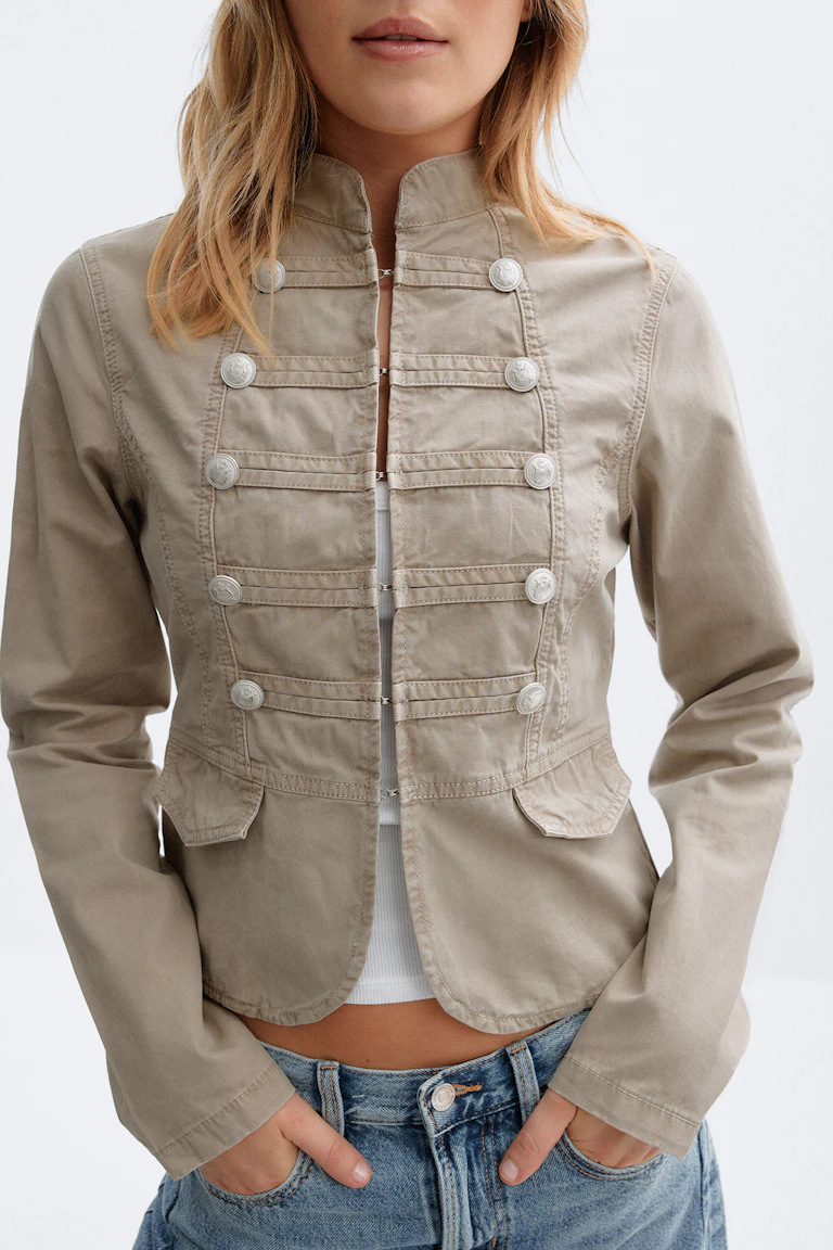 Hellbeige Kurzjacke im Military-Stil mit silbernen Knöpfen, kombiniert mit einem weißen gerippten Crop-Top und tief sitzenden Blue Jeans.