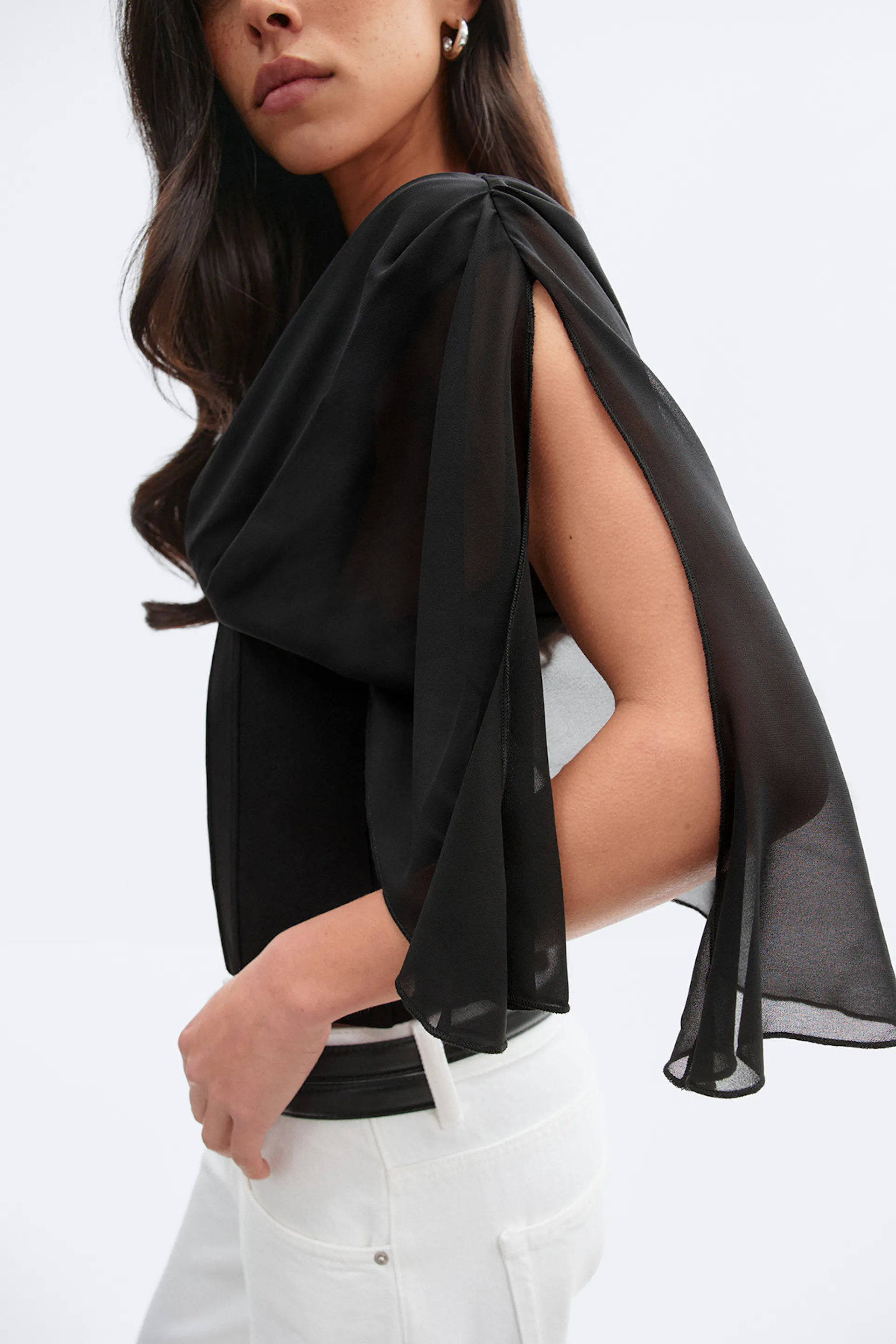 Schwarzes, durchsichtiges, drapiertes Cape-Top mit offenen, geschlitzten Ärmeln über einem taillierten, schwarzen Mieder, kombiniert mit weißen, hoch taillierten Jeans und einem schwarzen Gürtel.