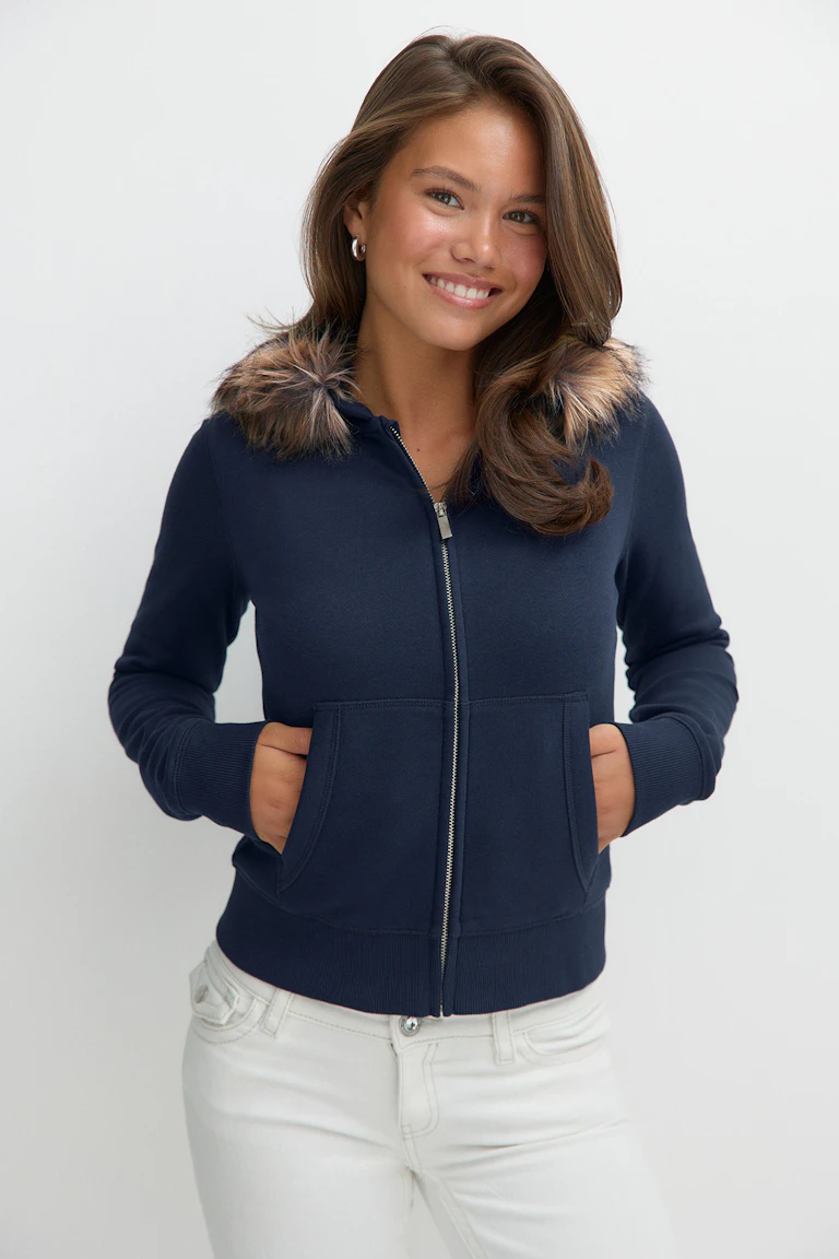 Mörkblå hoodie med dragkedja och kant av fuskpäls samt vita jeans.