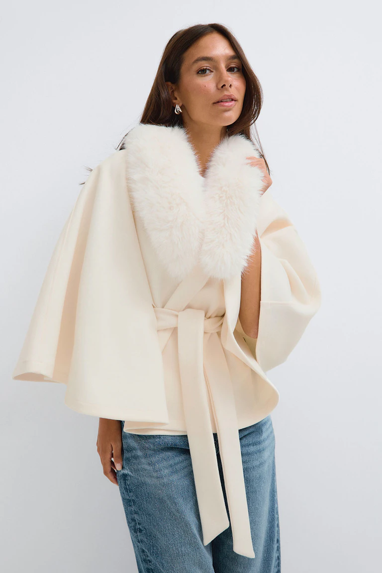 Cream belted cape with a faux-fur collar, paired with blue jeans.