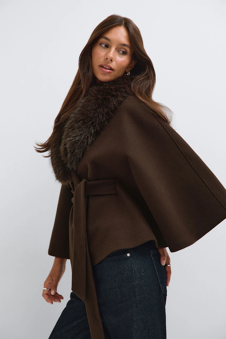 Model wearing a brown belted cape with a faux-fur collar, paired with dark jeans.