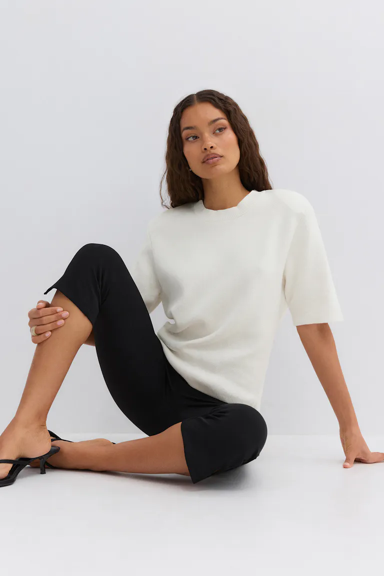 White short-sleeve crewneck knit top paired with black cropped slim-fit pants with small hem slits and black low-heeled mule sandals.