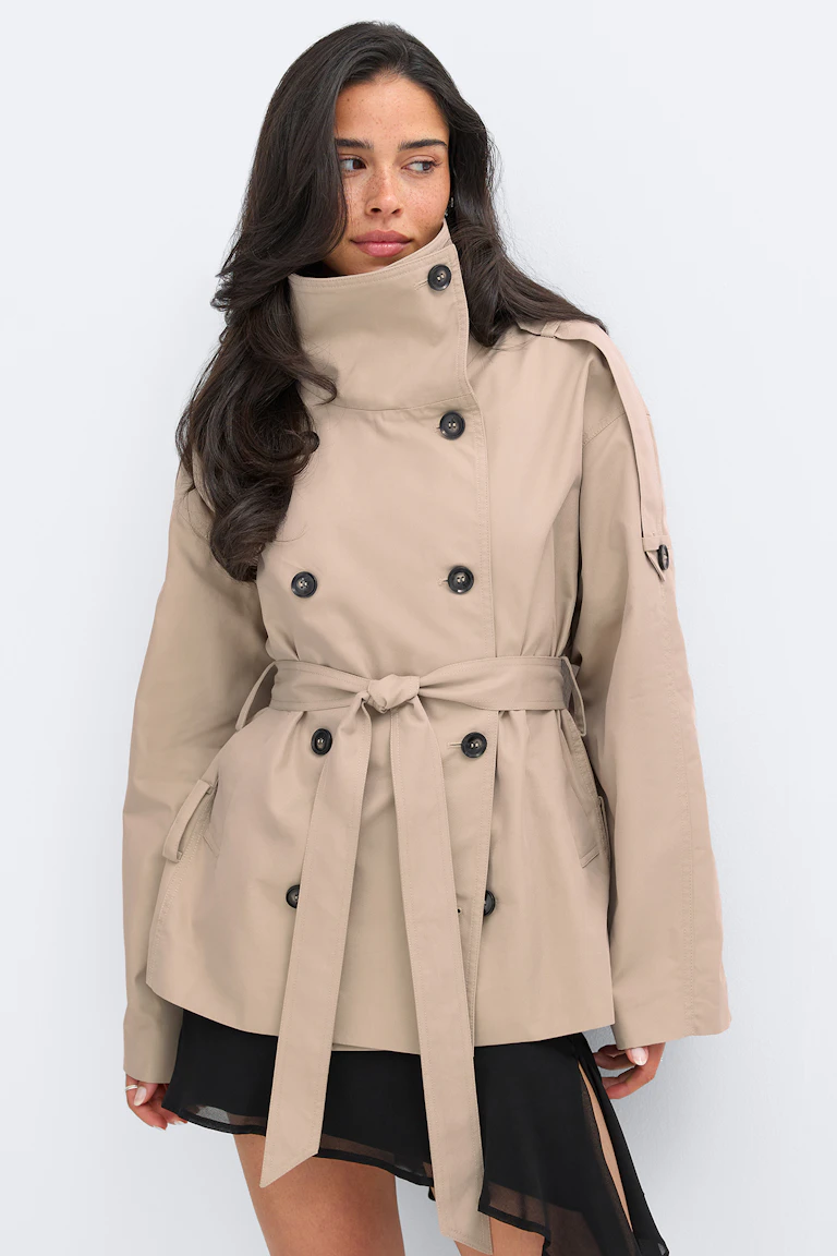 Beige trenchcoat med dubbelknäppning, hög ståkrage, svarta knappar, axelklaffar och ett knytbart midjebälte, stylad över en svart asymmetrisk kjol med genomskinlig fåll.