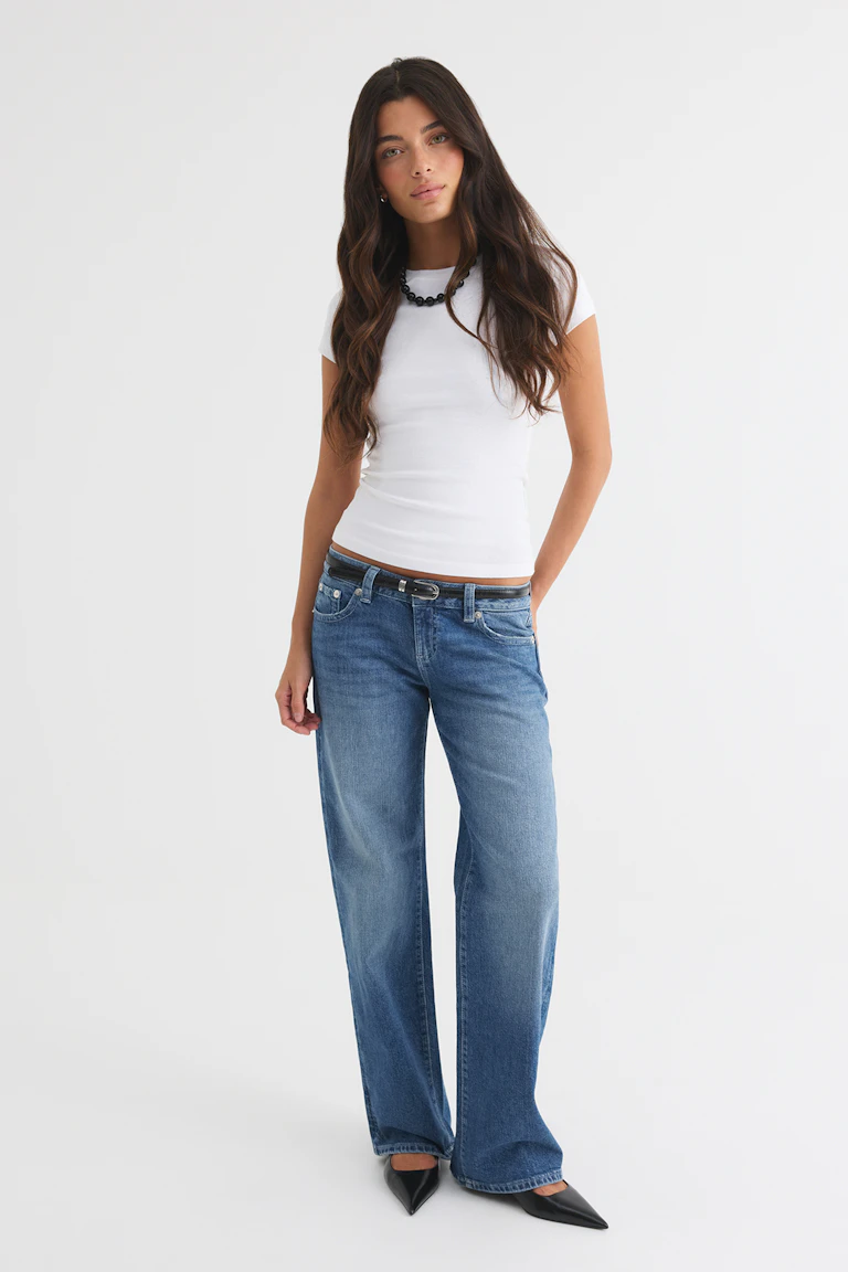 Een wit, nauwsluitend topje met korte mouwen, gecombineerd met een blauwe jeans met wijde pijpen, een zwarte riem, een zwarte kralenketting en zwarte puntige ballerina’s.