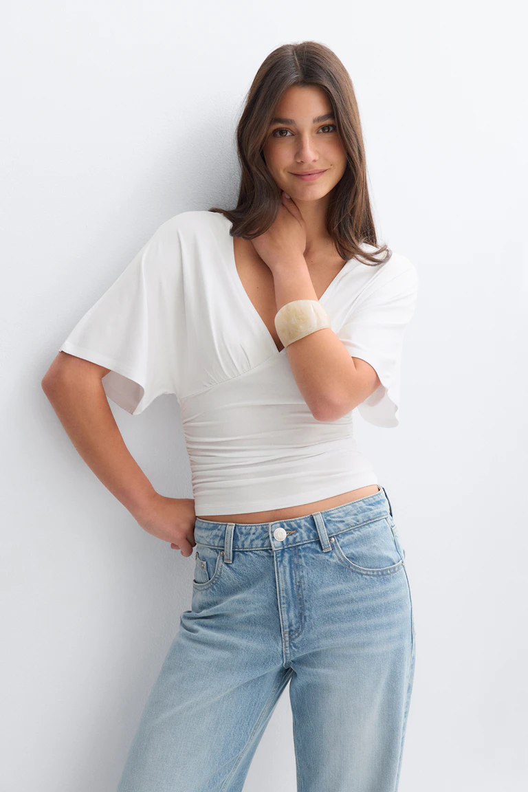 White V‑neck fitted top with short flutter sleeves, paired with light‑wash high‑rise jeans and a wide cream cuff bracelet.