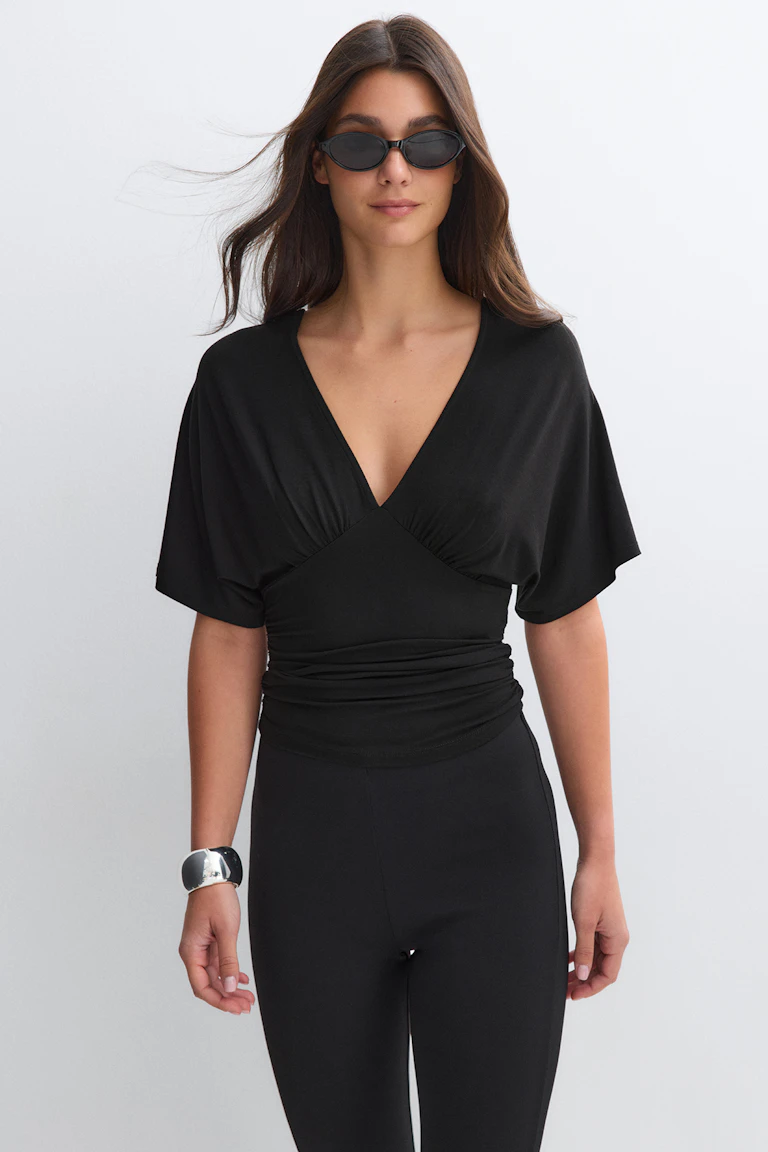 Schwarzes Wickeloberteil mit V-Ausschnitt, kurzen Flatterärmeln und geraffter Taille, taillierte schwarze Leggings mit hoher Taille, kleine schwarze ovale Sonnenbrille und ein breites silbernes Armband mit Manschetten.