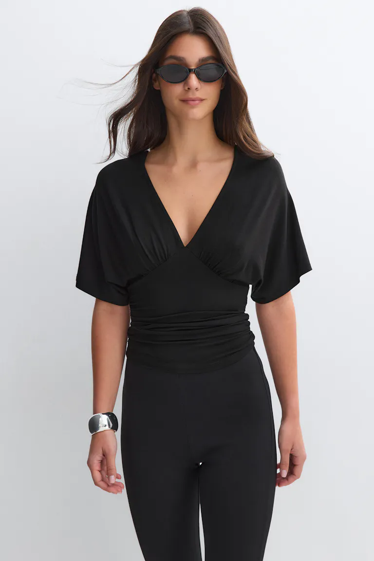 Black V-neck wrap-style top with short flutter sleeves and ruched waist, fitted high-waist black leggings, small black oval sunglasses, and a wide silver cuff bracelet.