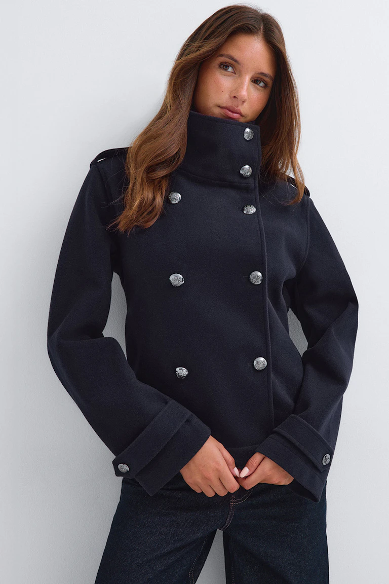 Structured dark navy double-breasted jacket with a high collar and silver button detailing, paired with dark jeans.
