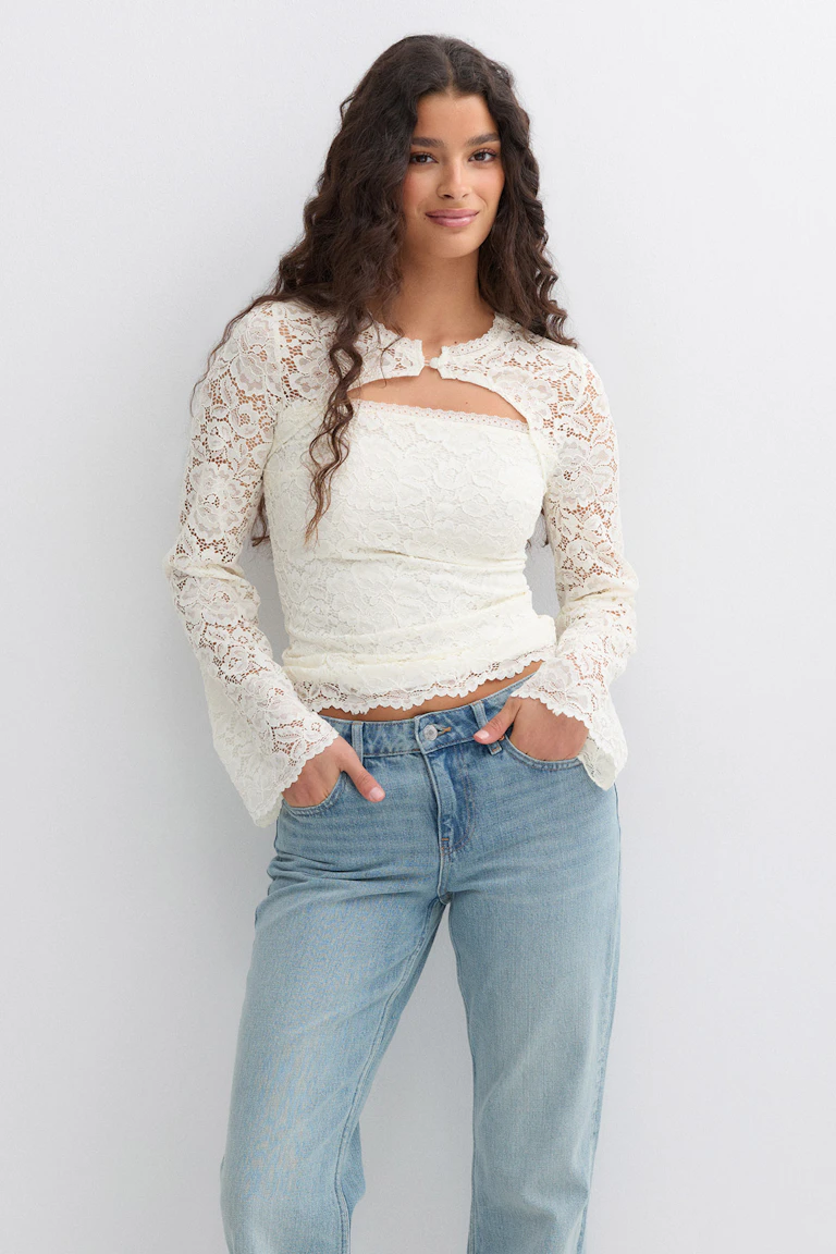 Långärmad bolero i elfenbensfärgad blommig spets med skulpterad kant, över en matchande axelbandslös camisole i spets, kombinerad med ljusa jeans med raka ben.