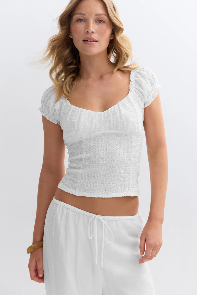 Weißes, strukturiertes, gesmoktes, kurzärmeliges Crop-Top mit gerüschtem Rundhalsausschnitt, kombiniert mit einer weißen Hose mit Kordelzug und einem Holz-Armreif.